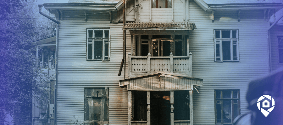 Weathered two-story house with boarded windows and a damaged porch, the kind of distressed property investors scout for deals.