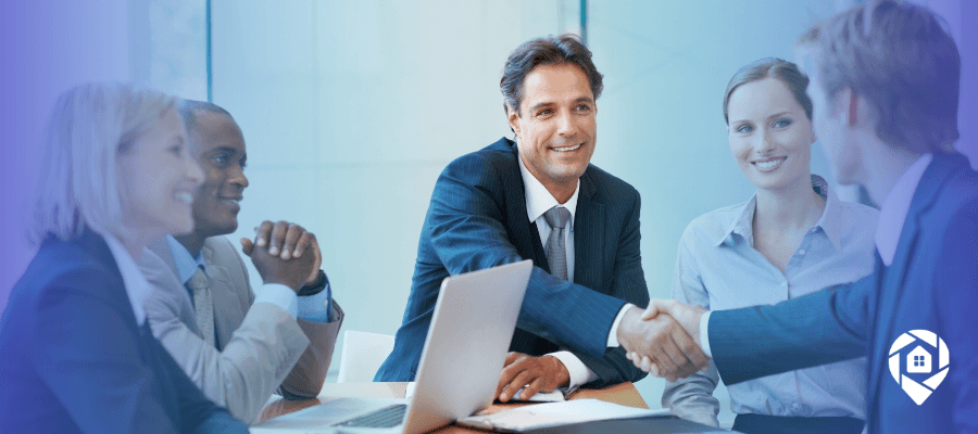 A team of business professionals at a conference table, two shaking hands over a laptop.